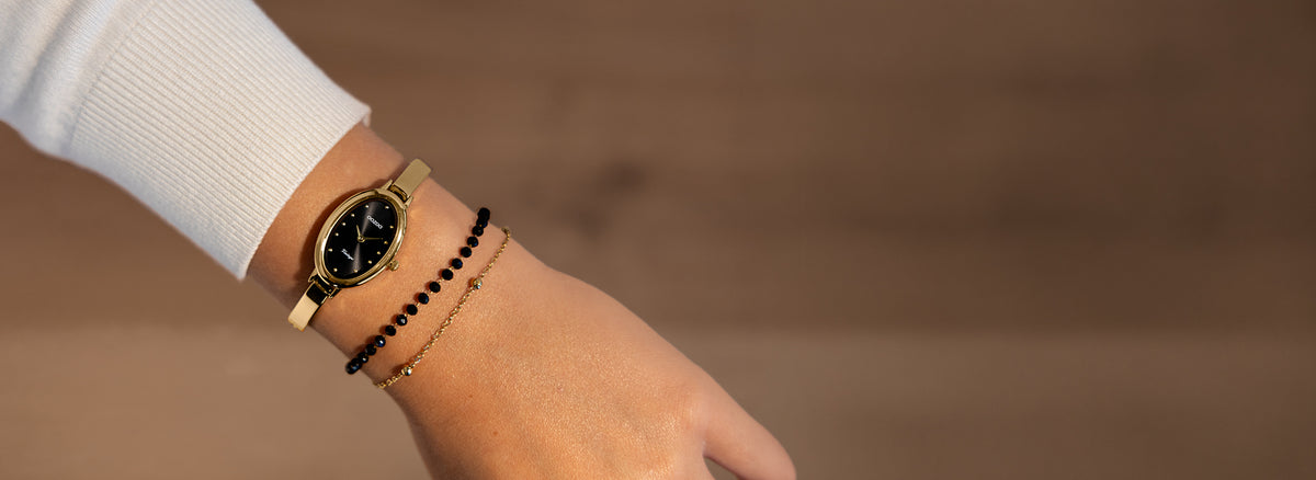 A close-up of a person's wrist wearing a gold-tone oval watch with a black dial, alongside two delicate black and gold bracelets, set against a warm, blurred background.
